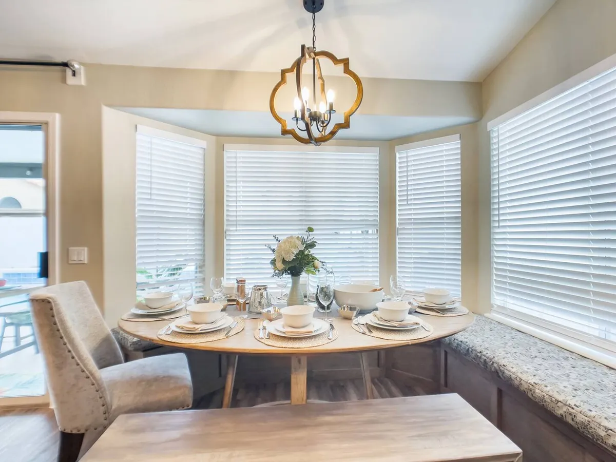 A beautifully set dining table with plates and glasses under a striking chandelier, accompanied by a cozy window seat and a fresh flower arrangement. The environment feels intimate and inviting, ideal for gathering, storytelling, and shared meals.