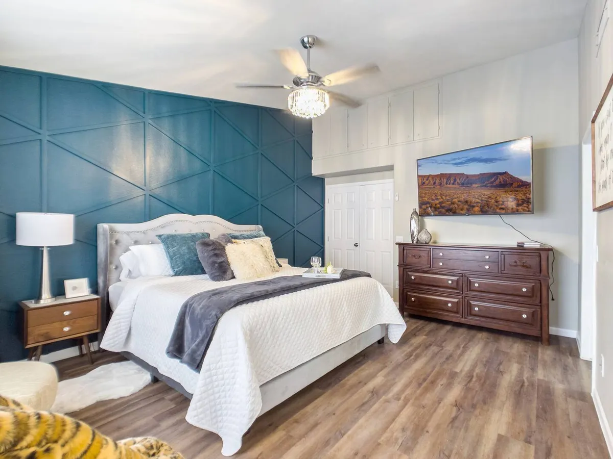 A tranquil bedroom featuring a bed with decorative pillows, a calming teal accent wall, a wooden dresser with a TV, and a nightstand with a lamp. The space is awash in soft natural light, creating a restful and inviting atmosphere ideal for unwinding.