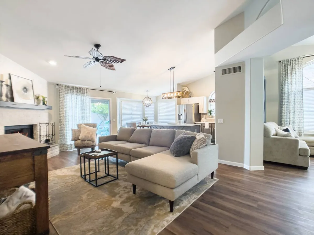 A spacious and light-filled living room featuring a plush sofa set, a central coffee table, a cozy fireplace, and a kitchen area visible in the background. Large windows bring in natural light, creating an inviting setting ideal for gatherings, relaxation, or enjoying a quiet moment by the fire.