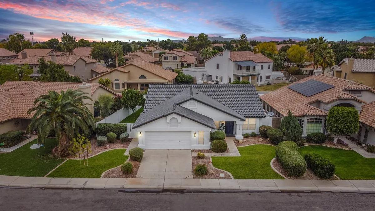 A wide shot of the home's front yard at sunset, with manicured landscaping, a long driveway, and the house's roof and inviting facade visible under a colorful sky. The scene is warm, approachable, and hints at arriving home after a day out.