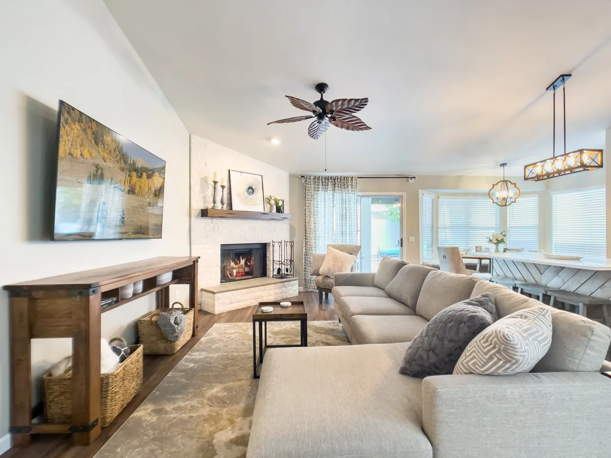 The image showcases a spacious, sunlit great room with a plush sectional sofa arranged around a cozy fireplace, a modern dining area with seating for a group, a stylish TV mounted above the mantle, and tasteful decor lining open shelves. The inviting layout exudes warmth and encourages relaxation or social gatherings, with plenty of space for family and friends to come together.