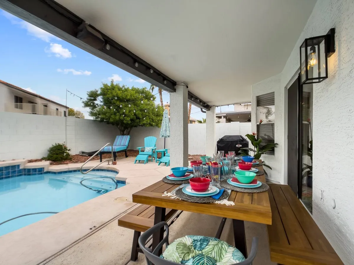 A cheerful backyard pool area with a large dining table set for a meal, colorful dishware, lounge chairs, and a grill. The setup radiates a festive, family-friendly vibe ideal for outdoor dining, celebrations, and poolside relaxation at home.