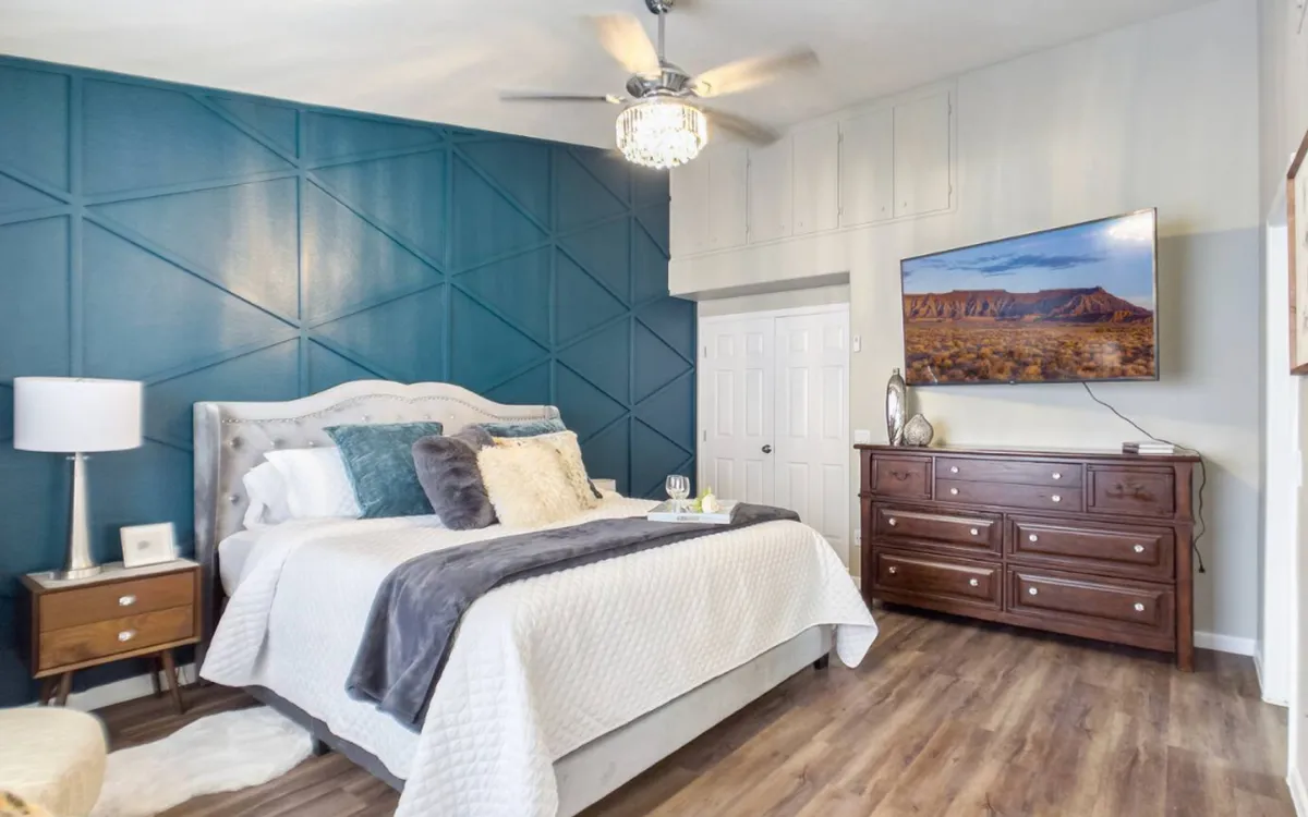 A stylish, tranquil bedroom with a comfortable bed adorned with decorative pillows, a modern ceiling fan, a wooden dresser supporting a TV, and an accent wall with a geometric pattern. Soft lighting and a cozy layout enhance the room's relaxing feel.