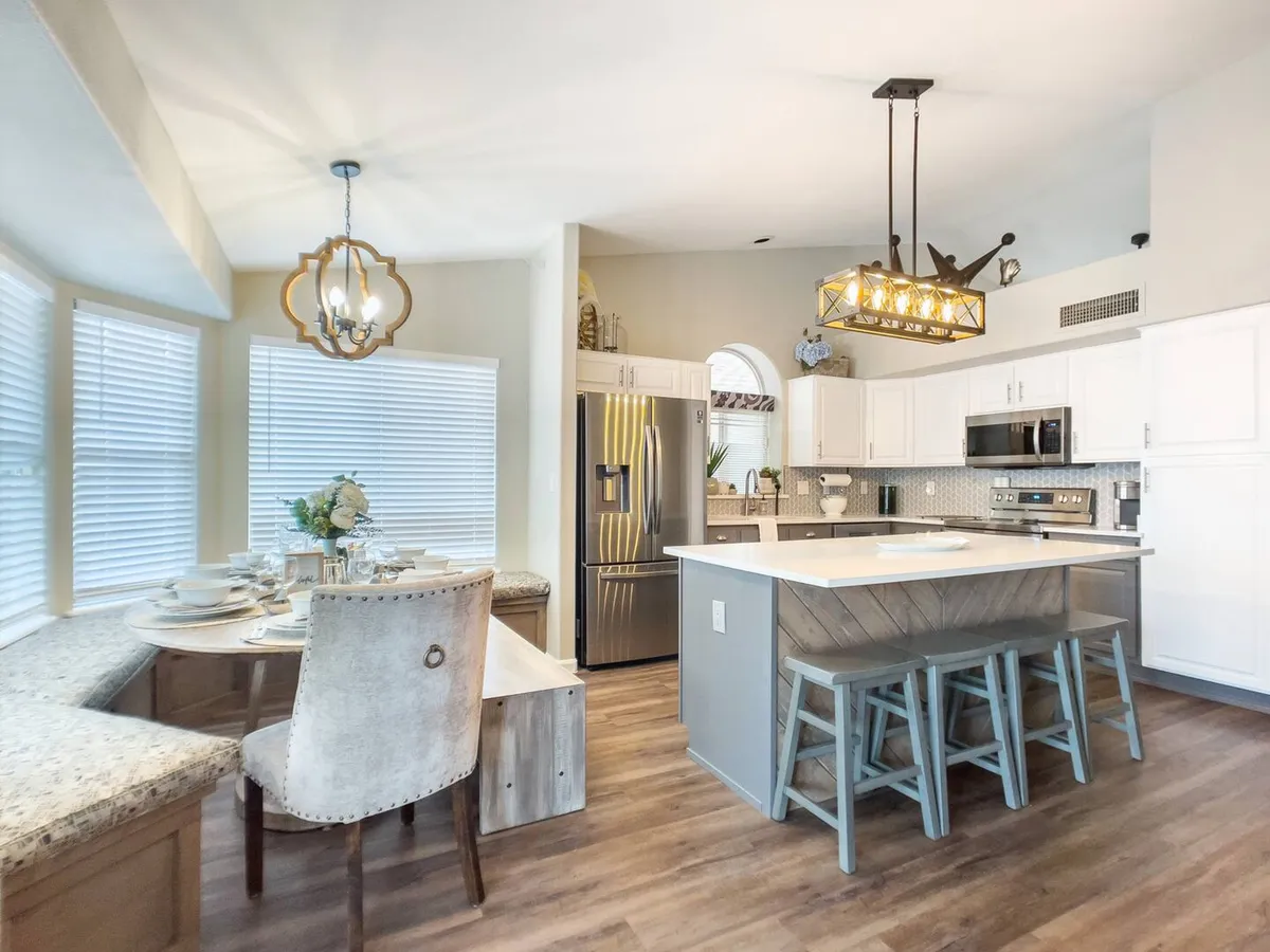 A spacious, light-filled kitchen and dining area with a central island, large windows, and visible modern appliances. The elegant table is set for a group, and the open layout encourages mingling and shared activities. Natural light streams in, enhancing the welcoming atmosphere.